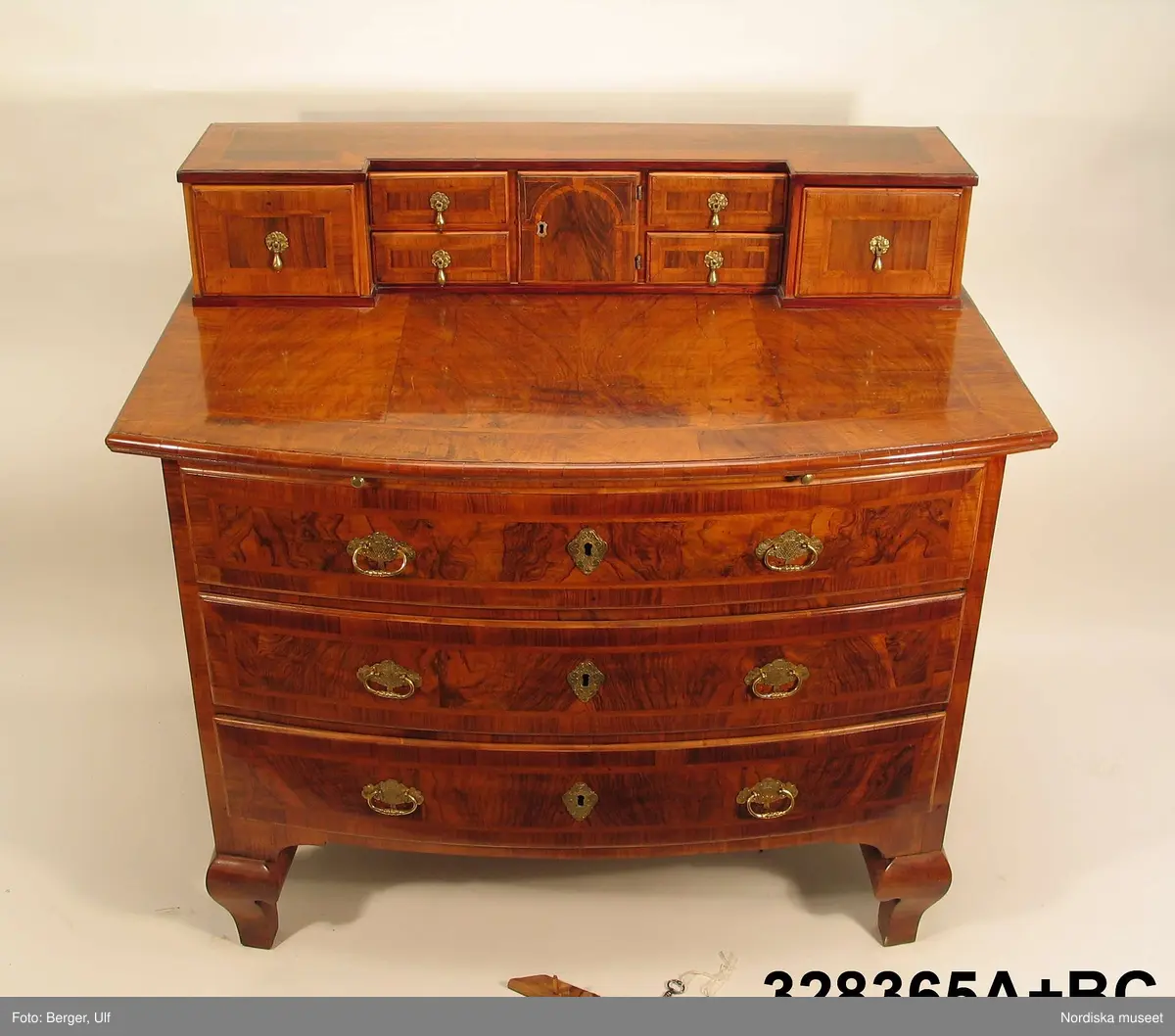 a+ = byrå
b = nyckel
c= nyckel
a+) Byrå, senbarock, Fredrik I:s tid, 1720-30-tal, trelådig, med uppsats, fanerad med valnöt. Raka sidor, svagt bågformad front och cabriolben. Utdragbar skrivskiva. Beslag av brännförgylld mässing. Stomme av furu, lådsidor och lådbottnar av ek.
Lådförstycken med fanerade fält av masurerad valnöt inramade av en dubbel fiskbensmönstrad åder och ytterst en fris. Utanpåliggande lådförstycken. Bygelhandtag med graverad dekor och sköldformade nyckelskyltar. (Delvis) senare lås, troligen 1800-tal.
Låg uppsats med två stora och fyra små lådor flankerande ett skåp, vars dörr har inlagt motiv av en valvbåge uppburen av kapitäl. Smålådorna med droppformade draghandtag.
På en av smålådornas baksidor mässingsplakett med text: "Tillhör R Elisabeth Thunberg / Intygas / G(?) Haglund".
b) Nyckel av järn till lådorna Längd 5,4 cm
c) Nyckel av järn till dörren på uppsatsen. 4,7 cm
/Anna Womack 2006-03-02