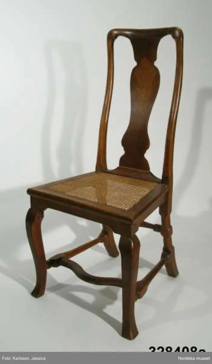 Stol, 6 st (a-f), England, 'Queen Anne', omkring 1720, av valnöt. Hög, balusterformad ryggbricka med inlagd (inkrusterad) dekor av ljusare träslag. Cabriolformade ben och svängt fotkryss med svarvade sidoslåar och bakre tvärslå. Sits med låg sarg, rottingflätad (rottingflätningen nygjord omkring 2002). Tillverkarinitialer inslagna på ben, ryggståndare och baksarg:
a) 'SH' (alt 'HS' ) samt 'IB'.
b) Rund stämpel: [kryss med dubbla streck] 
c) 'SH' (alt 'HS) samt 'IB'
d) 'SH' (alt HS') samt 'IB'
e) 'RH' (3 gånger) 
f) 'RH' (3 gånger),  'IB' (2 gånger) samt 'SH' (alt 'HS')

Jämför stol, inv nr 104.416 med initialerna 'RH' (enligt katalogkort). De engelska stolarnas märkning med tillverkarinitialer på t ex ben, sarg och ryggståndare visar på en rationalisering i tillverkningsprocessen. Initialerna är ibland placerade uppochnedvända vilket indikerar att de slogs in innan stolen sattes ihop. (se 'English furniture 1660-1714 - From Charles II to Queen Anne', Adam Bowett, Italien 2002, s. 264).
Anm: Flera stolar kompletterarade med delar av nytt trä, ilusade bitar. e) bakre tvärslå sekundärt förstärkt med skruv.
/Anna Womack 2008-09-25

Nordiska museet erbjöds 1992 att som testamentarisk gåva efter då framlidna Louise de Kiriline, Ontario, Kanada, ta emot en grupp sittmöbler bestående av två karmstolar och sex stolar. I samtal mellan arvinge till testator och dåvarande avdelningschefen Ingrid Bergman framgick att bakgrunden till gåvan var att del/delar av möblemanget redan skulle finnas i museets samlingar. En från Kanada översänd polaroidbild visade att en eller möjligtvis två stolar av denna typ, troligen tilllverkade i England omkr. 1720, förvärvats av museet redan 1905. Eftersom möbelgruppen skulle ge ett välkommet tillskott av engelska möbler till Charles Totties malmgård på Skansen tackade Nordiska museet ja till den erbjudna gåvan. Stolarna, som vid ankomsten till Sverige visade sig ha sekundära sitsar, har genom konservatorn Sören Lilja vid Skara museum åter försetts med rottingflätade sitsar av ursprunglig typ. Enligt testators anteckningar hade möblerna en gång tillhört den berömda romanförfattarinnan Sophie von Knorring (1797-1848). På sitt fäderne tillhörde hon ätten Zelow, som under 1700-talet haft stark förankring i Västergötland och även på hennes mans sida var det västgötska inslaget påfallande. De båda stolarna som redan tidigare tillhörde Nordiska museet, inv nr 104.415-104.416, inköptes 1905 från Göteborgs Historiska Museum dit de kommit från den kände samlaren Magister Nilsson i Skara. Dessa stolar är av samma typ, men olikheter i detaljer visar dock att de sannolikt ej ingått i samma möblemang. "Magister Nilsson", adjunkt vid Skara läroverk, som var en eldsjäl i Hazelius anda samlade kulturföremål från sin västgötska hembygd och de stora överensstämmelserna mellan dessa stolar och de stolar som nu förvärvas, visar att typen förekommit i Sverige under 1700-talet och kanske särskilt i de västra landskapen kring västerhavsporten Göteborg. Förvärvet behöver därför inte ses enbart relaterat till Tottieska malmgårdens möblering./Per Falck och Birgitta Martinius 2002.

Testator Louise de Kiriline Lawrence (född Flach, 1894-1992), var född i Sverige och växte upp på godset Svensksund på Vikbolandet i Östergötland. Efter att ha arbetat som röda korssyster under 1:a världskriget och i Ryssland efter revolutionen emigrerade hon till Kanada 1927. 