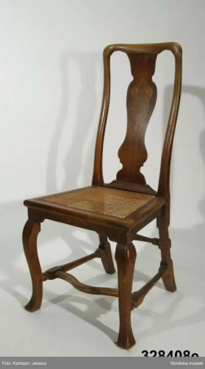Stol, 6 st (a-f), England, 'Queen Anne', omkring 1720, av valnöt. Hög, balusterformad ryggbricka med inlagd (inkrusterad) dekor av ljusare träslag. Cabriolformade ben och svängt fotkryss med svarvade sidoslåar och bakre tvärslå. Sits med låg sarg, rottingflätad (rottingflätningen nygjord omkring 2002). Tillverkarinitialer inslagna på ben, ryggståndare och baksarg:
a) 'SH' (alt 'HS' ) samt 'IB'.
b) Rund stämpel: [kryss med dubbla streck] 
c) 'SH' (alt 'HS) samt 'IB'
d) 'SH' (alt HS') samt 'IB'
e) 'RH' (3 gånger) 
f) 'RH' (3 gånger),  'IB' (2 gånger) samt 'SH' (alt 'HS')

Jämför stol, inv nr 104.416 med initialerna 'RH' (enligt katalogkort). De engelska stolarnas märkning med tillverkarinitialer på t ex ben, sarg och ryggståndare visar på en rationalisering i tillverkningsprocessen. Initialerna är ibland placerade uppochnedvända vilket indikerar att de slogs in innan stolen sattes ihop. (se 'English furniture 1660-1714 - From Charles II to Queen Anne', Adam Bowett, Italien 2002, s. 264).
Anm: Flera stolar kompletterarade med delar av nytt trä, ilusade bitar. e) bakre tvärslå sekundärt förstärkt med skruv.
/Anna Womack 2008-09-25

Nordiska museet erbjöds 1992 att som testamentarisk gåva efter då framlidna Louise de Kiriline, Ontario, Kanada, ta emot en grupp sittmöbler bestående av två karmstolar och sex stolar. I samtal mellan arvinge till testator och dåvarande avdelningschefen Ingrid Bergman framgick att bakgrunden till gåvan var att del/delar av möblemanget redan skulle finnas i museets samlingar. En från Kanada översänd polaroidbild visade att en eller möjligtvis två stolar av denna typ, troligen tilllverkade i England omkr. 1720, förvärvats av museet redan 1905. Eftersom möbelgruppen skulle ge ett välkommet tillskott av engelska möbler till Charles Totties malmgård på Skansen tackade Nordiska museet ja till den erbjudna gåvan. Stolarna, som vid ankomsten till Sverige visade sig ha sekundära sitsar, har genom konservatorn Sören Lilja vid Skara museum åter försetts med rottingflätade sitsar av ursprunglig typ. Enligt testators anteckningar hade möblerna en gång tillhört den berömda romanförfattarinnan Sophie von Knorring (1797-1848). På sitt fäderne tillhörde hon ätten Zelow, som under 1700-talet haft stark förankring i Västergötland och även på hennes mans sida var det västgötska inslaget påfallande. De båda stolarna som redan tidigare tillhörde Nordiska museet, inv nr 104.415-104.416, inköptes 1905 från Göteborgs Historiska Museum dit de kommit från den kände samlaren Magister Nilsson i Skara. Dessa stolar är av samma typ, men olikheter i detaljer visar dock att de sannolikt ej ingått i samma möblemang. "Magister Nilsson", adjunkt vid Skara läroverk, som var en eldsjäl i Hazelius anda samlade kulturföremål från sin västgötska hembygd och de stora överensstämmelserna mellan dessa stolar och de stolar som nu förvärvas, visar att typen förekommit i Sverige under 1700-talet och kanske särskilt i de västra landskapen kring västerhavsporten Göteborg. Förvärvet behöver därför inte ses enbart relaterat till Tottieska malmgårdens möblering./Per Falck och Birgitta Martinius 2002.

Testator Louise de Kiriline Lawrence (född Flach, 1894-1992), var född i Sverige och växte upp på godset Svensksund på Vikbolandet i Östergötland. Efter att ha arbetat som röda korssyster under 1:a världskriget och i Ryssland efter revolutionen emigrerade hon till Kanada 1927. 