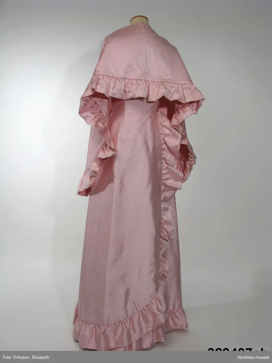 Aftonklänning med Sjal.
a) Aftonklänning av blekt rosa shantungsiden. hellång modell med baddräktsringat åtsittande liv och vid kjol. Helskuren med vid, båtformig urringning som är djupare bak och löper i ett med smala axelband. Bystinsnitt samt längsgående insyningar över byst , mitt fram samt på ryggstycket. Framflyttade sidsömmar. Omlottskuren baktill med mjukt rundad bakvåd. Efter denna från midjan och ner runt kjolen löper en volang av dubbelt tyg. Blixtlås i vänster sida.
Klolen fodrad med styvt nylonnät. Infodringar efter livets urringning, nedtill på kjolen och efter hela omlottvåden. Ryggstycket dubblerat på ett intrikat sätt. 
b) Schal av samma tyg som klänningen, långsmal modell med ett rundat hörn i vardera änden och med volang efter ena långsidan.
Bars med specialkomponerat smycke av glaspärlor.
Komponerad av Gunilla Pontén till Karin Hyldgaard-Jensen, vinnare av Fröken Sverige-titeln 1962. Vinnaren tävlade senare under året om titeln "Miss International Beauty" i Long Beach, Californien. Där kom Karin med till semifinalen. Hela resegarderoben komponerades av Gunilla Pontén. Den kompletterades med konfektionsplagg som också valdes ut av G.P. Till detta kom också en "folkdräkt" som var tung och varm och inte särskilt populär hos Karin. 
 Se inv.nr 329.405-329.407 samt tillbehör 329.585-329.587
/Ingrid Roos2009-06-23