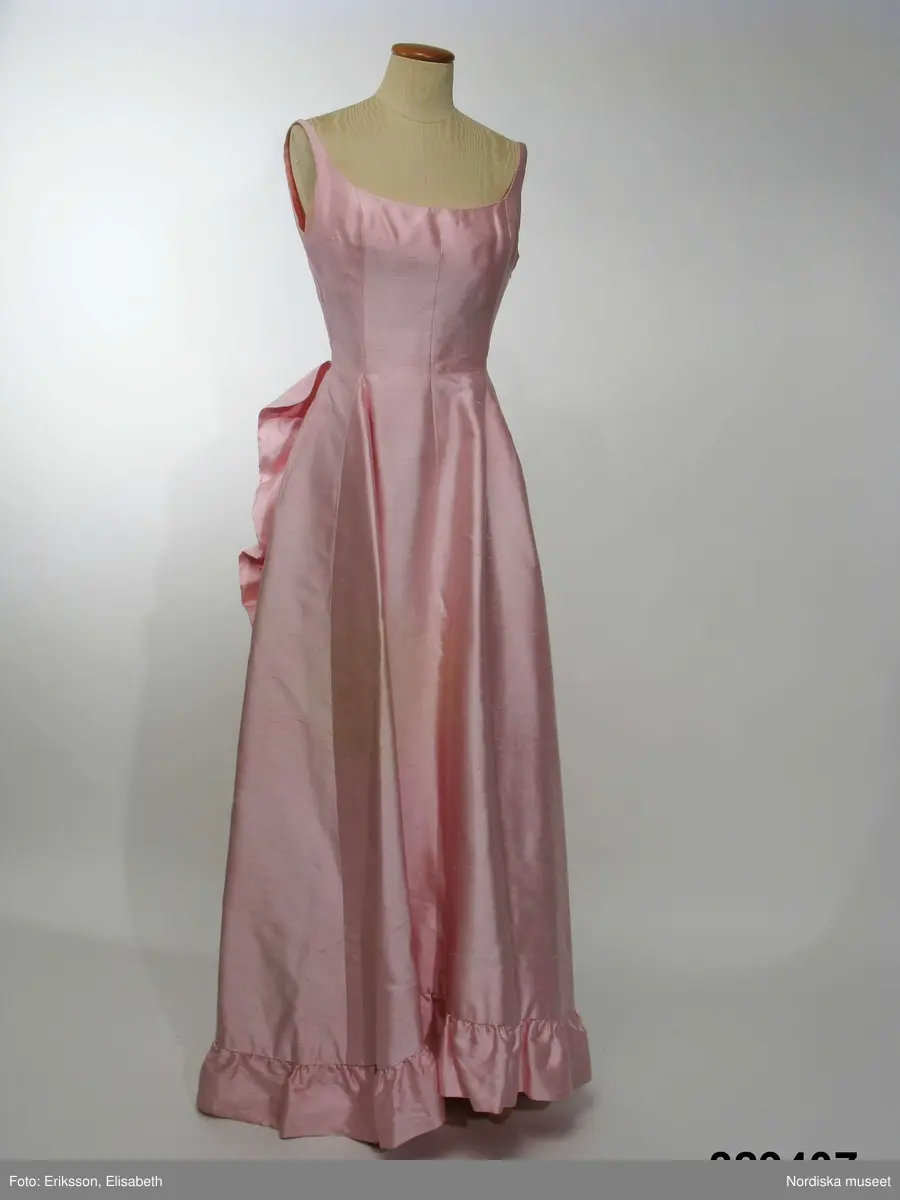 Aftonklänning med Sjal.
a) Aftonklänning av blekt rosa shantungsiden. hellång modell med baddräktsringat åtsittande liv och vid kjol. Helskuren med vid, båtformig urringning som är djupare bak och löper i ett med smala axelband. Bystinsnitt samt längsgående insyningar över byst , mitt fram samt på ryggstycket. Framflyttade sidsömmar. Omlottskuren baktill med mjukt rundad bakvåd. Efter denna från midjan och ner runt kjolen löper en volang av dubbelt tyg. Blixtlås i vänster sida.
Klolen fodrad med styvt nylonnät. Infodringar efter livets urringning, nedtill på kjolen och efter hela omlottvåden. Ryggstycket dubblerat på ett intrikat sätt. 
b) Schal av samma tyg som klänningen, långsmal modell med ett rundat hörn i vardera änden och med volang efter ena långsidan.
Bars med specialkomponerat smycke av glaspärlor.
Komponerad av Gunilla Pontén till Karin Hyldgaard-Jensen, vinnare av Fröken Sverige-titeln 1962. Vinnaren tävlade senare under året om titeln "Miss International Beauty" i Long Beach, Californien. Där kom Karin med till semifinalen. Hela resegarderoben komponerades av Gunilla Pontén. Den kompletterades med konfektionsplagg som också valdes ut av G.P. Till detta kom också en "folkdräkt" som var tung och varm och inte särskilt populär hos Karin. 
 Se inv.nr 329.405-329.407 samt tillbehör 329.585-329.587
/Ingrid Roos2009-06-23