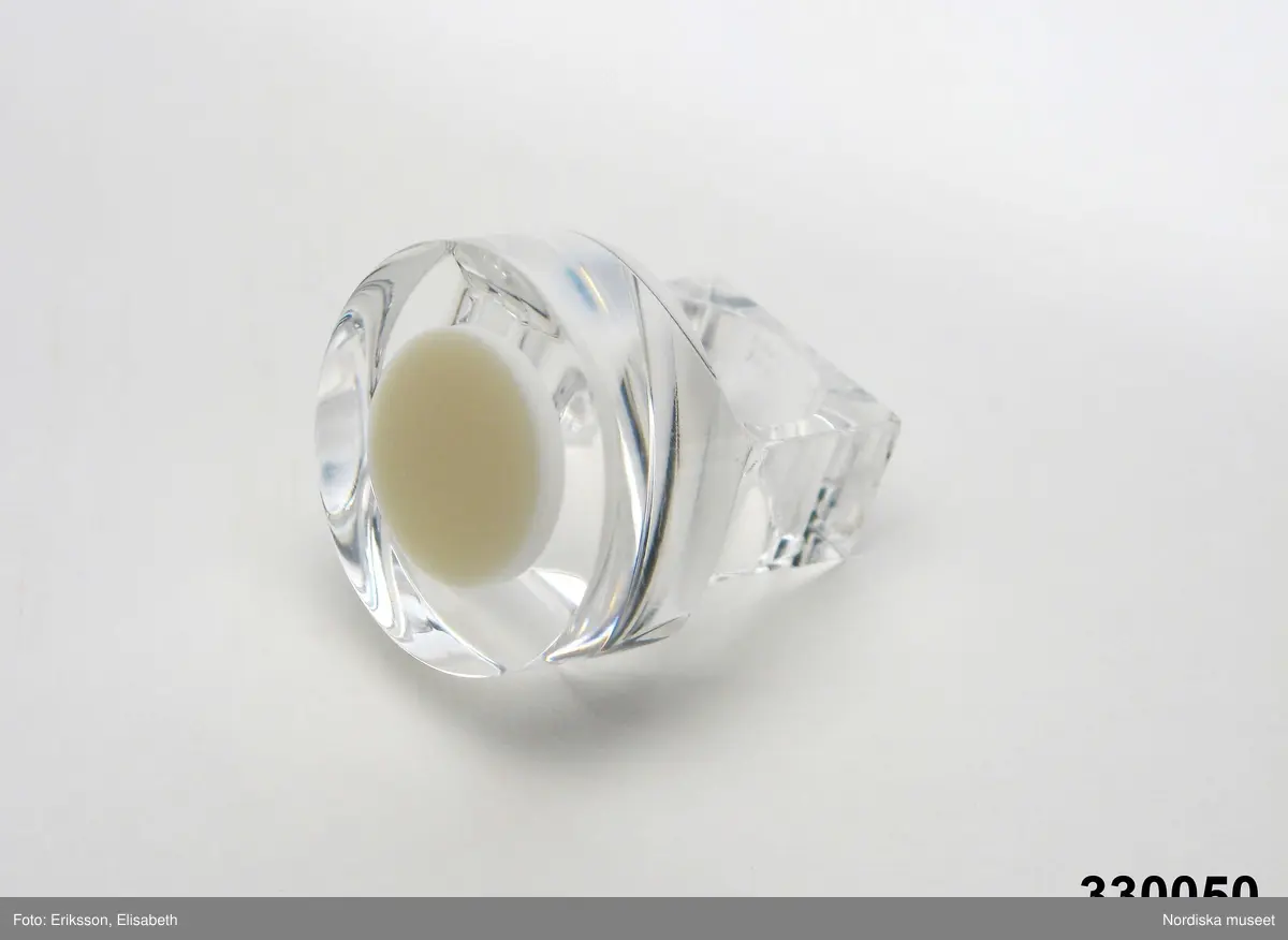 Ring av transparent akrylplast med konformig inläggning av vit plast. Cirkelrunt framstycke, skena med yttre kvadratisk form. Designad av silversmeden Siv Lagerström under 1960-talets senare del. Tillverkad av Gravyrverken i Sollentuna (råämne som kapades och frästes ur) och Siv Lagerström (urborrning för fyllning av färgad Castolite, en form av gjutplast). Inskription på skenans bakre del "Siv" samt "MADE IN SWEDEN". 
/Ingrid Roos 2010-04-07