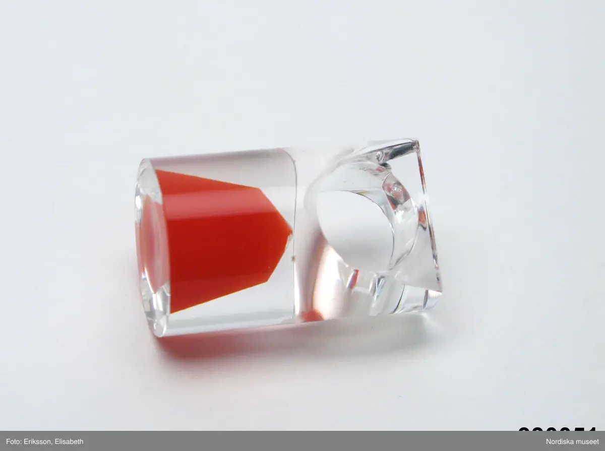 Ring av transparent akrylplast med konformig inläggning av röd plast. Cirkelrunt framstycke, skena med yttre kvadratisk form. Designad av silversmeden Siv Lagerström under 1960-talets senare del. Tillverkad av Gravyrverken i Sollentuna (råämne som kapades och fräster ur) och Siv Lagerström (urborrning för fyllning av färgad Castolite, en form av gjutplast). Inskription på skenans bakre del "Siv" i skrivstil.
/Ingrid Roos 2010-04-07