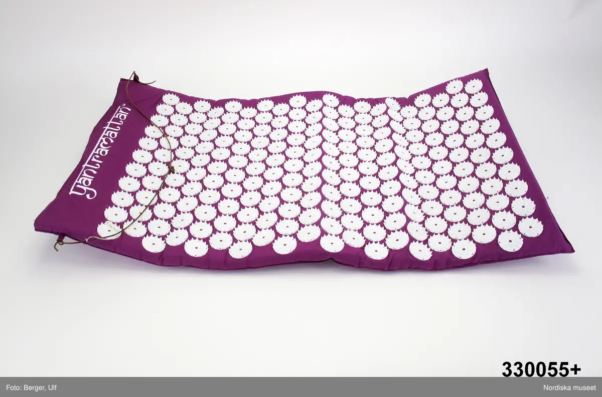 Spikmattan Yantra. Storlek medium. Mattan består av ett rektangulärt tygstycke. Dubbelt tyg med öppning i ena kortänden, fästs ihop med kardborreband. På mattans ena sida finns 210 vita "plastblommor"/rundlar. Varje blomma består av 42 bladspetsar/piggar. Det blir sammanlagt 8 820 piggar. Rundlarna är tillverkade av 100% giftfri ABS-plast och tyget av 100% bomull. På andra sidan finns en tryckt "yantrasymbol" i vitt. Inuti uttagbar skumplast, 78x46 cm. Vid öppningen fäst i öglor en smal läderrem som "låser" mattan när man viker den i tre delar vid förvaring. Inuti mattan vit etikett med  text "ABS PLASTIC" och tvättanvisningar (symboler) samt "CE".
/Lena Kättström Höök och Elin Nystrand von Unge 2010-04-13