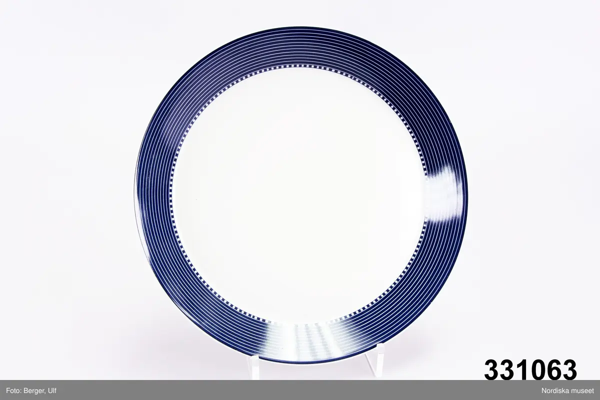 Mattallrik, modell Teema, stengods. Tillverkad av Arabia, design Kaj Frank. Vit och blå tallrik, med mörkblå hög utåtlutande kep med smala vita linjer samt vid mötet mellan kep och spegel dekor av vita och blå kvadrater. Vit  odekorerad spegel. Inget bräm.
Stämplad under tallriken.

/Cecilia Wallquist 2011-07-19