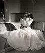 Franska avdelningen vid Nordiska Kompaniet 1941. Kvinnlig modell sitter i en soffa iförd hellång klänning med blusliv med kort puffärm, liten krage och knäppning. Kjolen är i flera lager och vid. Skärp i avvikande färg.