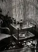 Interiör med tropikmöbler av bambu, rotting och glas från varuhuset Nordiska Kompaniet i Stockholm 1942. På en serveringsvagn står karaff och glas, på den undre hyllan ligger  en bok. Intill serveringsvagnen en stol och gröna växter, i bakgrunden en jalusi som silar ljuset.