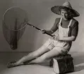 Sommarmode från varuhuset Nordiska Kompaniet i Stockholm 1944. En kvinnlig modell iklädd korta hängselbyxor, tröja, stråhatt och sandaler sitter med en fiskehåv i handen. Vid hennes sida står en väska av flätat näver.