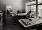 Inredningsavdelningens ritkontor på varuhuset Nordiska Kompaniet i Stockholm 1947. Två män sitter vid varsitt skrivbord och arbetar med ritningar. Ljuset faller in genom ett stort fönster. Bakom dem ser man genom en dörröppning in i ytterligare ett kontorsrum där skjortklädda män arbetar.