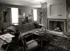 Interiör med vardagsrumsmöbler från varuhuset Nordiska Kompaniet i Stockholm 1947. Framför den öppna spisen en soffa, fåtölj och dukat kaffebord samt ett brickbord med keramiköverdel. på golvet en mönstrad ryamatta. Stormönstrade tapeter med bladrankor.