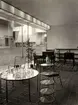 Interiör från Stockholmsutställningen 1930 med möbler från Nordiska Kompaniet. Ett runt bord och ett avlastningsbord i flera etager, båda av stålrör med glasskivor. På bordet står en grupp slipade glas och karaffer. Intill står två karmstolar av stålrör med stoppade dynor. I bakgrunden ses en del av utställningsområdet
