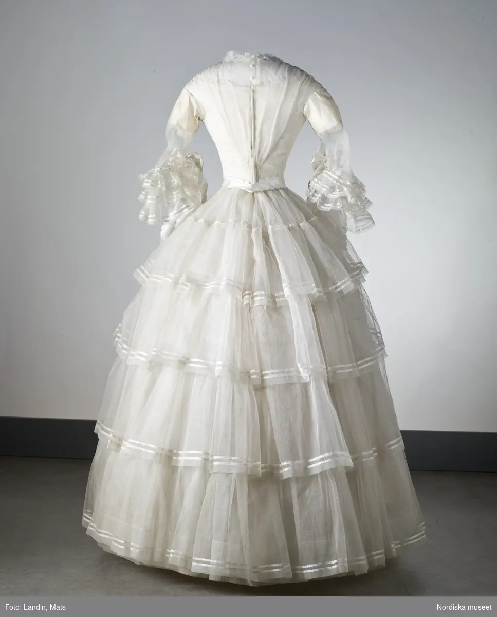 Brudklänning, liv med fastsydd kjol, vit tarlatan, garnerad med vita sidenband, rosetter. Klänningen har ett löst skärp av vit tarlatan.