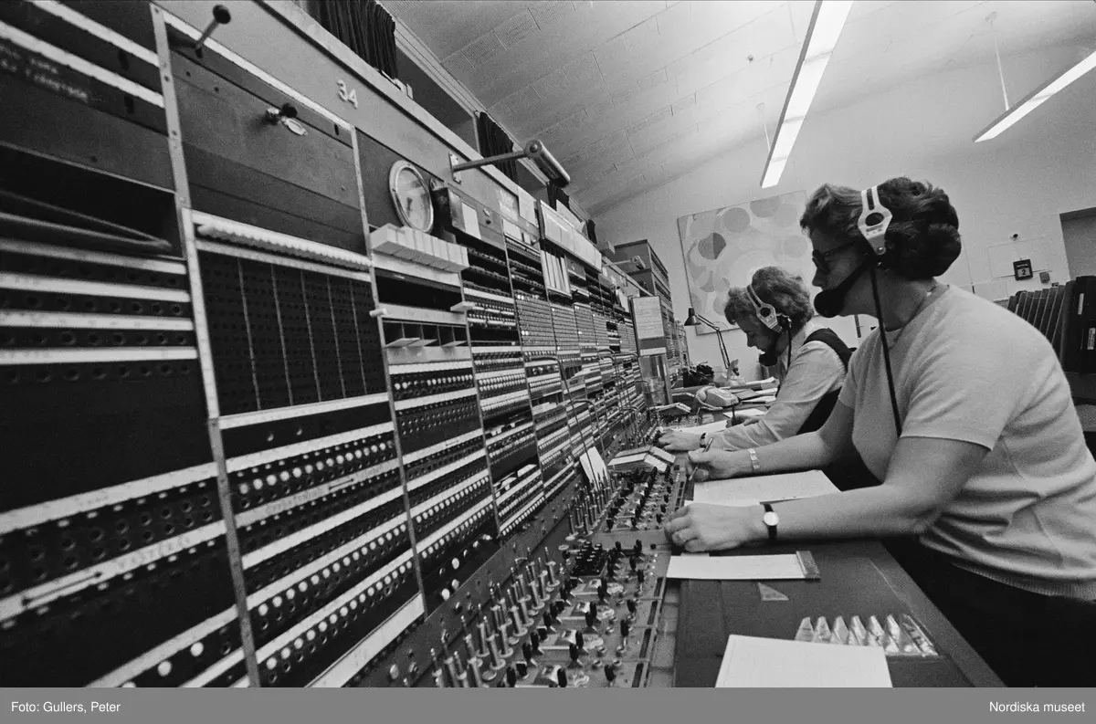 Telefonväxel. Några kvinnor arbetar med hörlurar och mikrofon framför en panel med knappar och reglage.