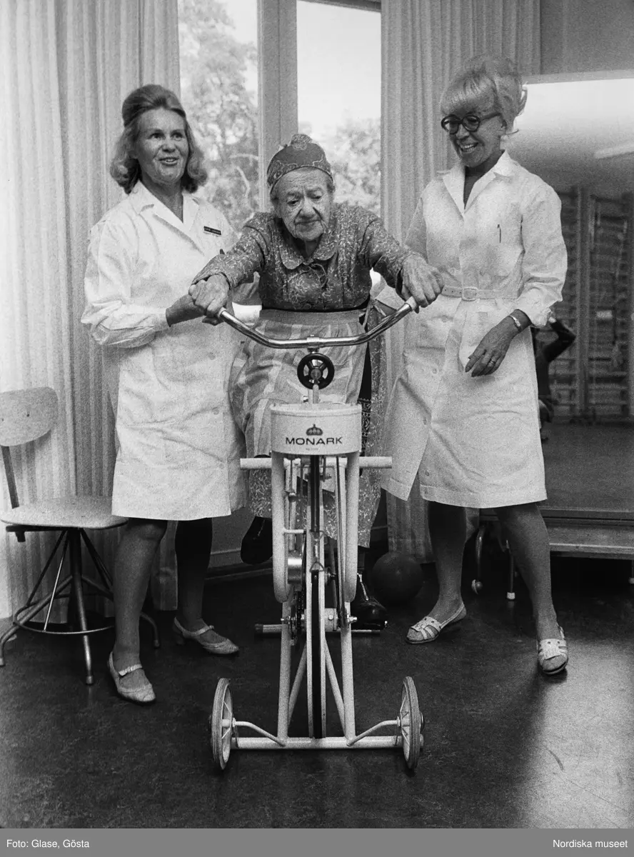 En gammal kvinna på motionscykel av märket Monark flankerad av vårdpersonal i vita rockar.