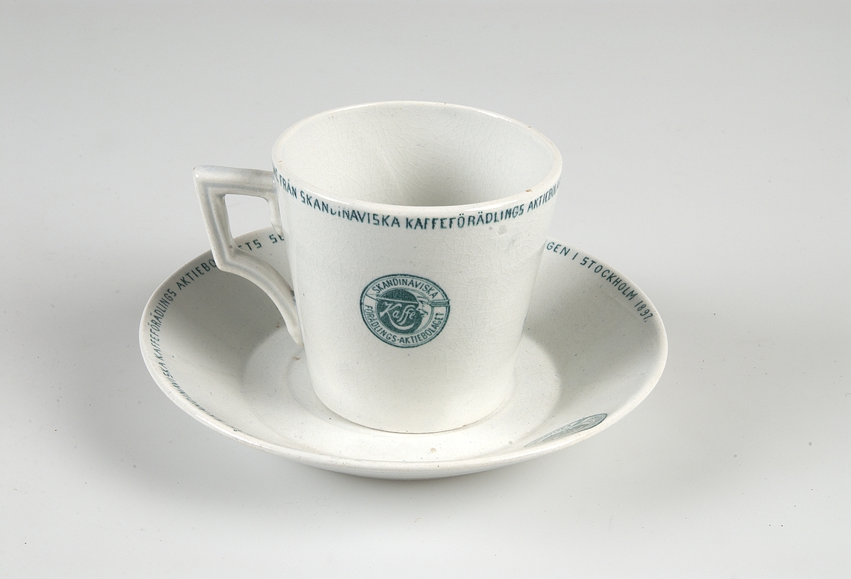Kaffekopp med fat, vitglaserade med grön dekor. Runt kanten: "Minne från skandinaviska kaffeförädlings aktiebolags serveringspaviljong å utställningen i Stockholm 1897". Samma text på fatet. Grön tryckt stämpel: "GUSTAFSBERG".