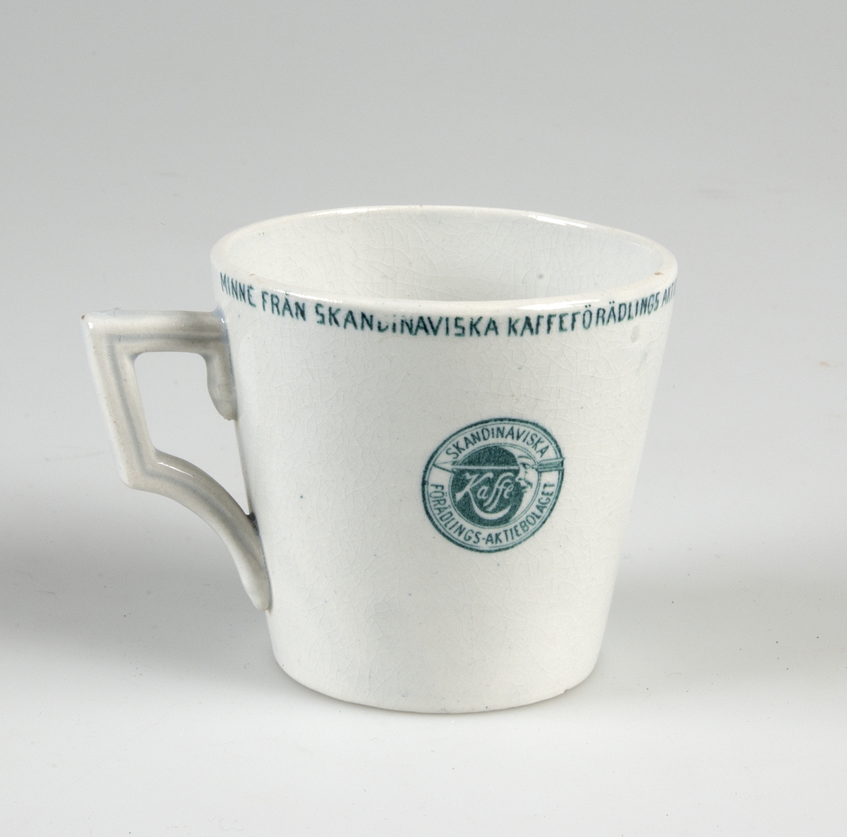 Kaffekopp med fat, vitglaserade med grön dekor. Runt kanten: "Minne från skandinaviska kaffeförädlings aktiebolags serveringspaviljong å utställningen i Stockholm 1897". Samma text på fatet. Grön tryckt stämpel: "GUSTAFSBERG".