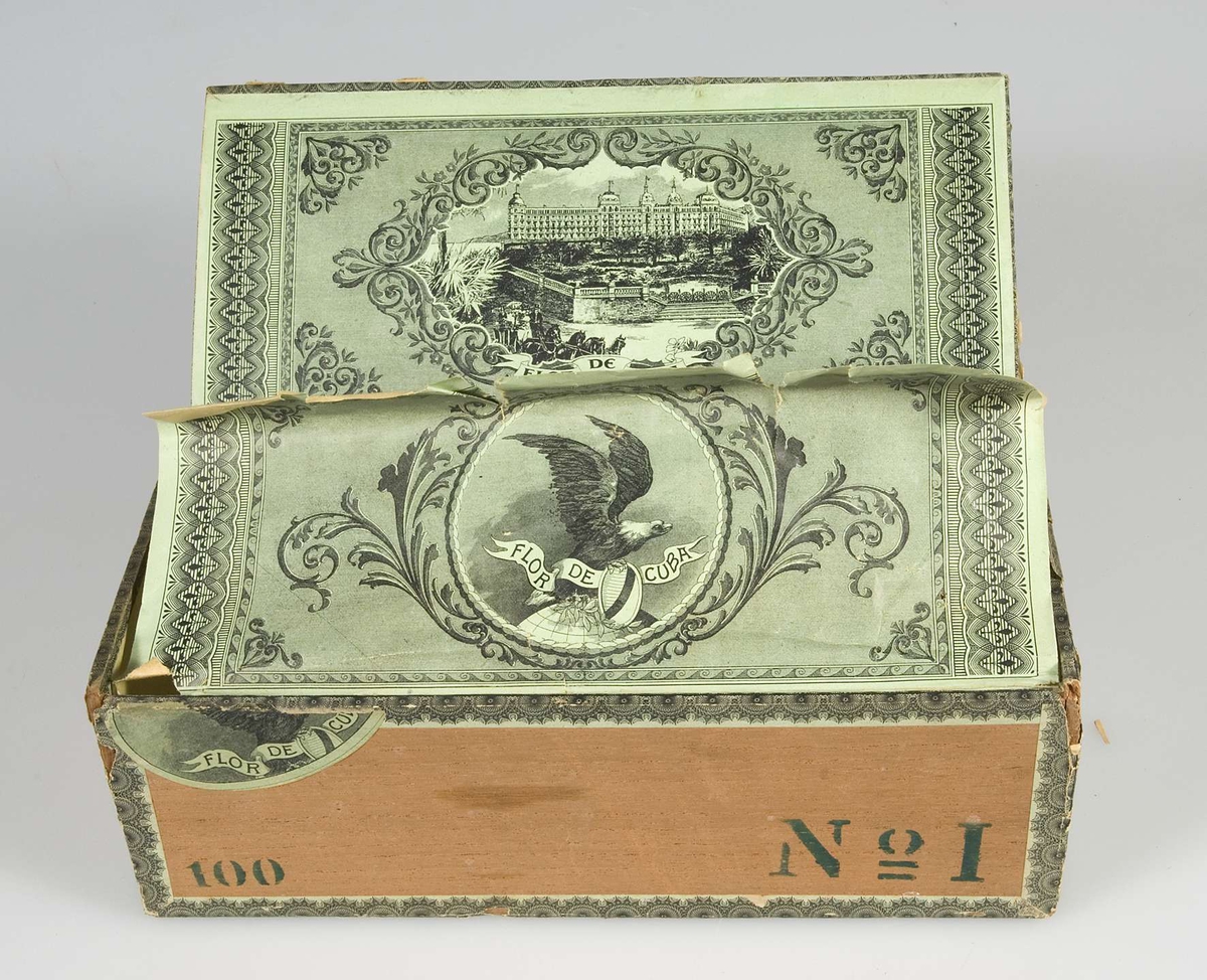 Papperslister i grönt och svart. Text på lockets pappersetiketter: Flor de Cuba. På locket en stämpel med texten: Havanna Magasinet Stockholm; Flor de Cuba, samt en örn. Blyertsanteckningar på locket: Till julgran. På framsidan i grönt tryck: 100, No 1. Fodrad med grönt papper. På lockets insida ett byggnadsmotiv i svart, på skyddsbladet en örn i svart.
