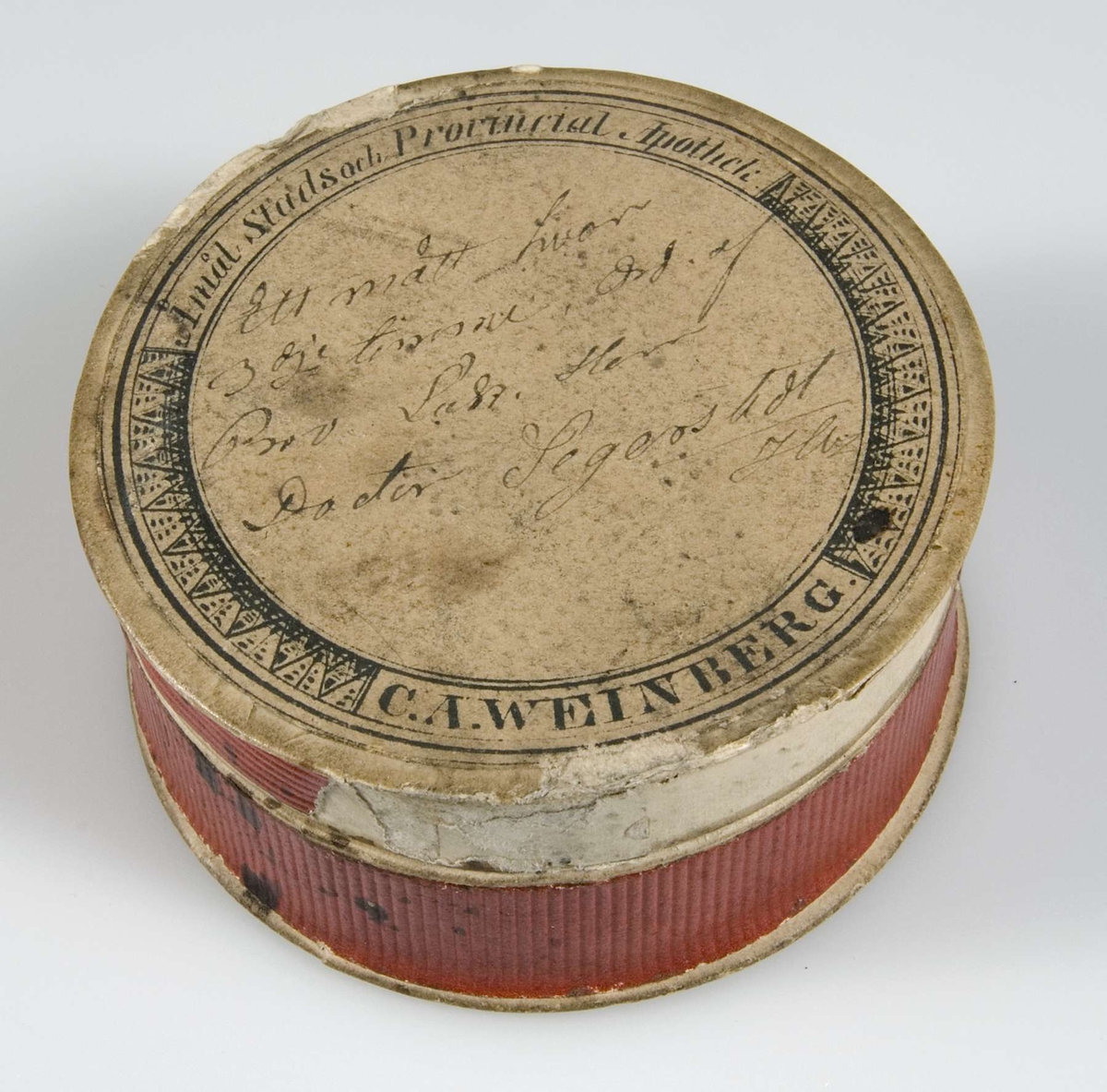 Ask av papp, röda sidor. Vit etikett med svart text "Åmål stads och Provincial Apotek C. A. Weinberg", med bläck "Ett mått hvar 3 dje timme, Ord. af Prov. Las. Herr Doktorn Segerstedt".

