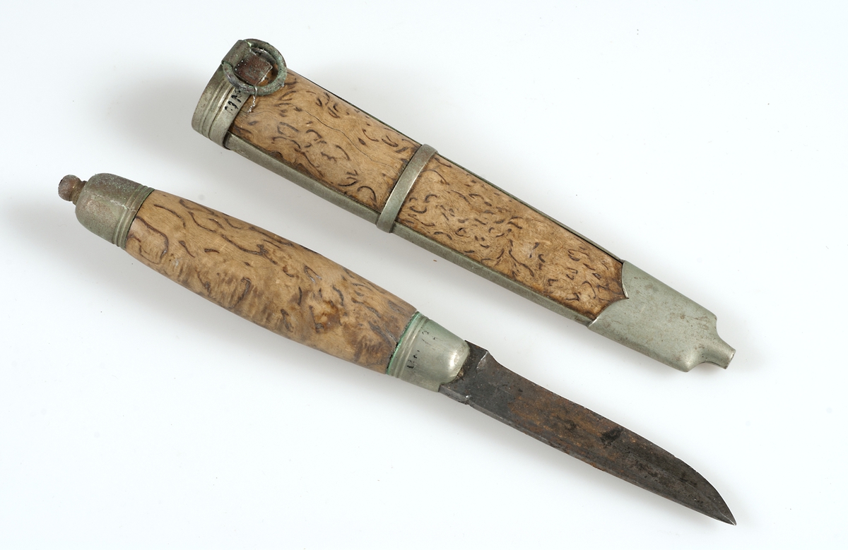 Kniv med slida av trä.
a) Kniv: skaft av masurbjörk, beslag av vitmetall, blad av stål. 
b) Slida: av samma träslag som knivskaftet, vitmetallbeslag, ring fästad i överkantens beslag.   
Kniven har använts vid korgbindning.