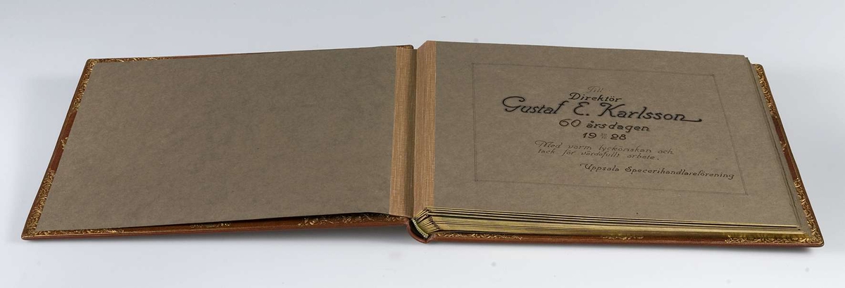Album, inbundet i helfranskt band i brunt läder med stämplade ramverk och ornament i guld. På pärmens framsida spegelmonogram, komponerat av bokstäverna GEK (Gustaf E. Karlsson). Guldsnitt runt om, blad av mörkgrå kartong. Frontespis med textad tillägnan: "Till Direktör Gustaf  E. Karlsson 60-årsdagen 19 30/10 28. Med varm lyckönskan och tack för värdefullt arbete. Uppsala Specerihandlarförening". Albumet innehåller fotografier av Gustaf E. Karlsson, av hans speceriaffär, av styrelsen i Uppsala Specerihandlarförening samt av föreningens hedersledamöter och medlemmar. Albumet på pärmens baksida stämplat: "Levins Bokbinderi. Uppsala Bokband C W Bergman". Mapp, klädd med mörkbrunt läder. 