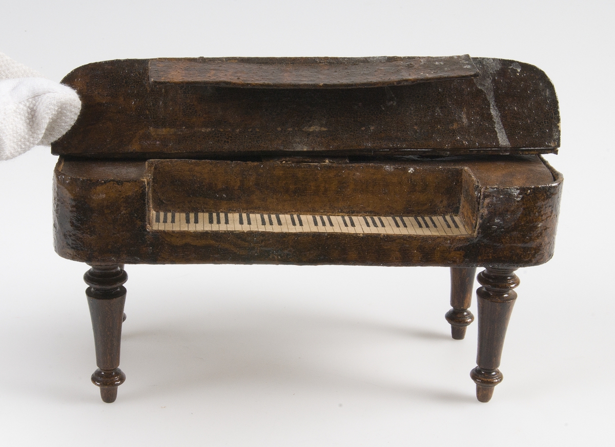 Leksak, taffel av mörkbrun och gulflammig ädelträimitation. Klaviatur av papper. På undersidan står med bläck: i Rde 20 öre. 