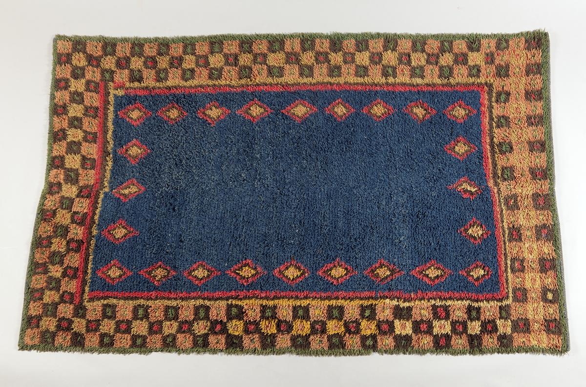 Prydnadsrya i två våder, hopsydda kant i kant. Botten i korndräll med varp av blått s-tvinnat lingarn 11 tr/cm och inslag av mörkbrun s-spunnen ull 7½ inslag/cm. Nockor av en tråd tvåtrådigt z-tvinnat hårdtvinnat ullgarn, knutna med giordesknut över 4 varptrådar med 4 varptrådar mellan nockorna och 4 inslag mellan nockraderna. Nockorna syns ej på baksidan.

Mönster: Stort blått mittfält avgränsat mot bården av en röd, en grönbrun och en gul smal linje. På det blå mittfältet, utmed sidorna romber av rött, grönbrunt och gult. Bred bård av schackrutor i grönbrunt, gult och rosagult. Gröna, röda och gula prickar i rutorna. Ytterst smal grön rand.