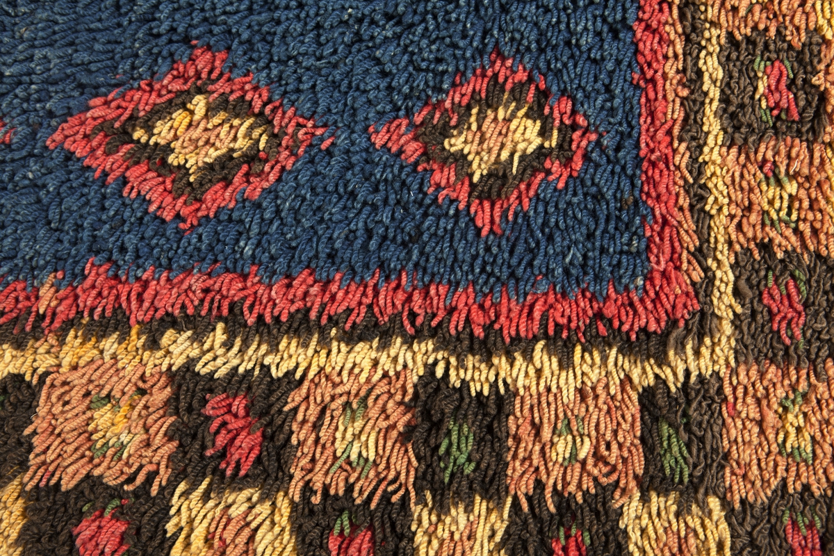 Prydnadsrya i två våder, hopsydda kant i kant. Botten i korndräll med varp av blått s-tvinnat lingarn 11 tr/cm och inslag av mörkbrun s-spunnen ull 7½ inslag/cm. Nockor av en tråd tvåtrådigt z-tvinnat hårdtvinnat ullgarn, knutna med giordesknut över 4 varptrådar med 4 varptrådar mellan nockorna och 4 inslag mellan nockraderna. Nockorna syns ej på baksidan.

Mönster: Stort blått mittfält avgränsat mot bården av en röd, en grönbrun och en gul smal linje. På det blå mittfältet, utmed sidorna romber av rött, grönbrunt och gult. Bred bård av schackrutor i grönbrunt, gult och rosagult. Gröna, röda och gula prickar i rutorna. Ytterst smal grön rand.