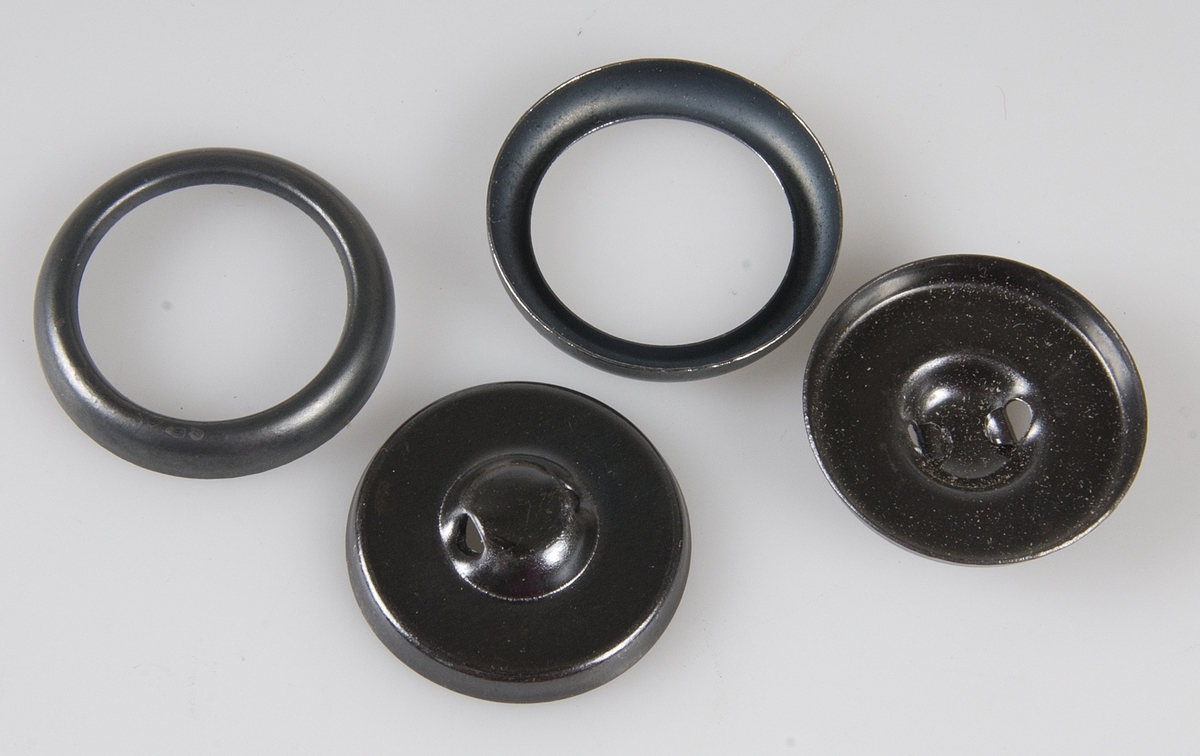 Originalförpackning, kartong av oblekt papp indelad i tre fack med 100 knappformar i tre delar, dels av svart metall, ringar och knappar med kanal, dels runda ljusbruna pappskivor. Knappformarnas diameter ca 2 cm. Ovanpå locket papperstejp, på sidan etikett med text: HAGEX H & S knappformar DUBBELMONTAGE (utan inlägg) 5 gross 36" svart Art. 2 (med kanal).
