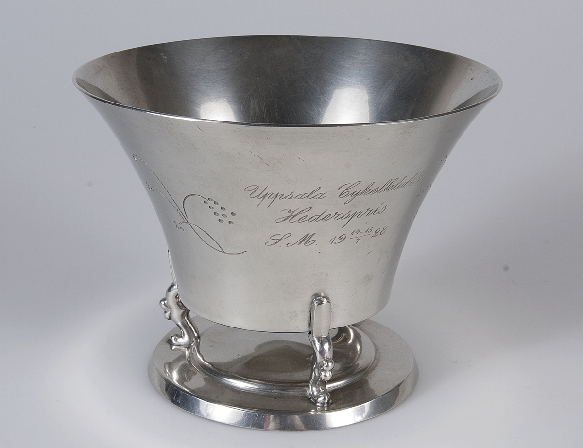 Vid skål av tenn med tre tassar som förbinder skålen med en bottenplatta. En blomsterslinga på var sida om inskriptionen. Ingraverat: Uppsala Cykelklubbs Hederspris, SM 14-15/7 1928.