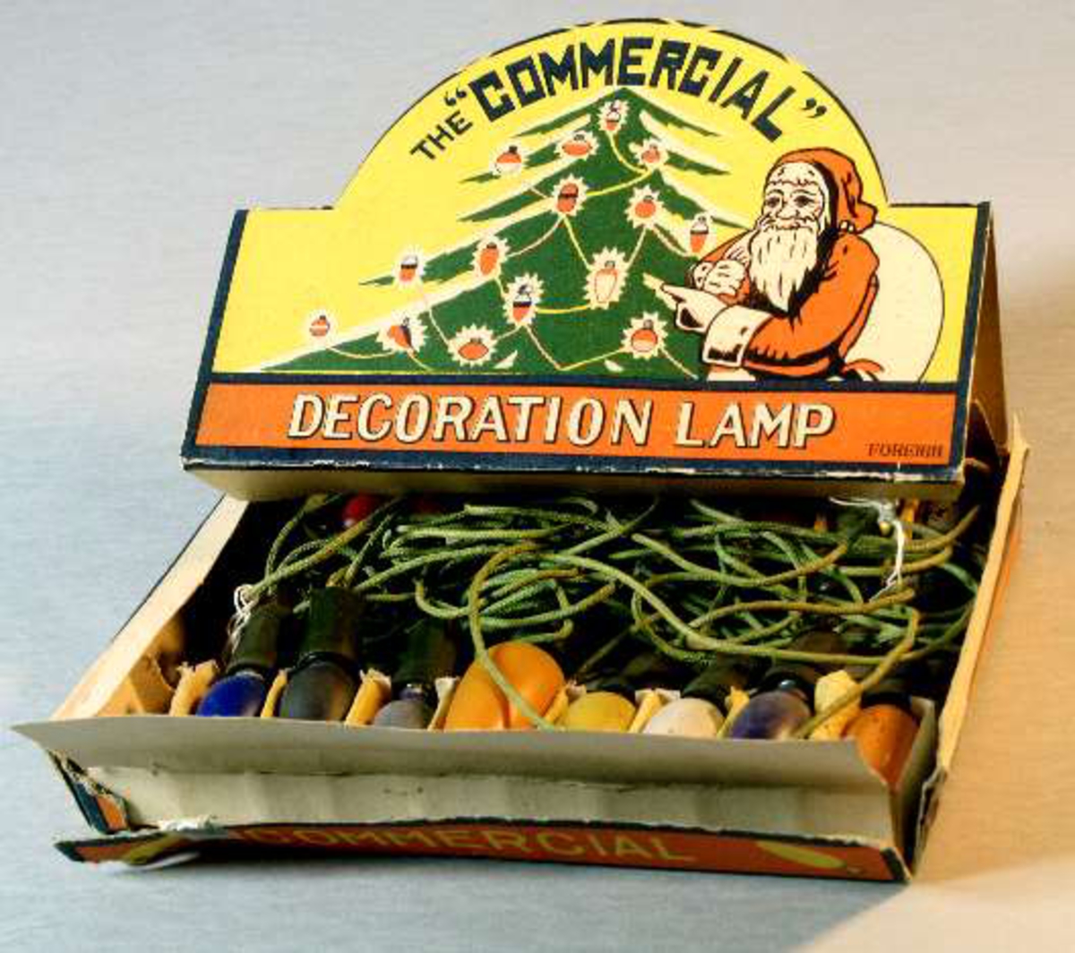 Pappask innehållande kulört elektrisk julgransbelynning, 16 små äggformade lampor av glas i blått, gult, vitt , rött, grönt och orange , i hållare av grön plast och med sladdar klädda med grön tråd. Några lampor är större än de andra och troligen utbytta. På askens lock bild av tomte och julgran i rött, gult, grönt och vitt och text: The Commersial Decoration lamp. Foreign.