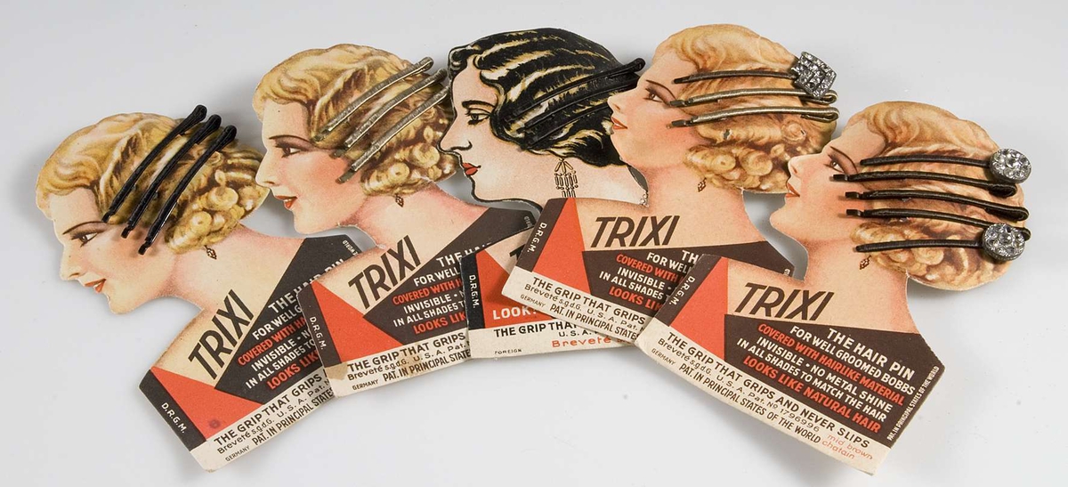 48 förpackningar med två - tre smala klädda hårspännen i varje förpackning. Förpackningarna formade till kvinnoansikte. Tryckt text: TRIXI THE HAIR PIN. Förpackningarna förvaras i blåvit ask. Tillverkade i USA. På baksidan av varje förpackning stämplat 30 resp. 40 öre. Två förpackningar har hårspännen av strass.
