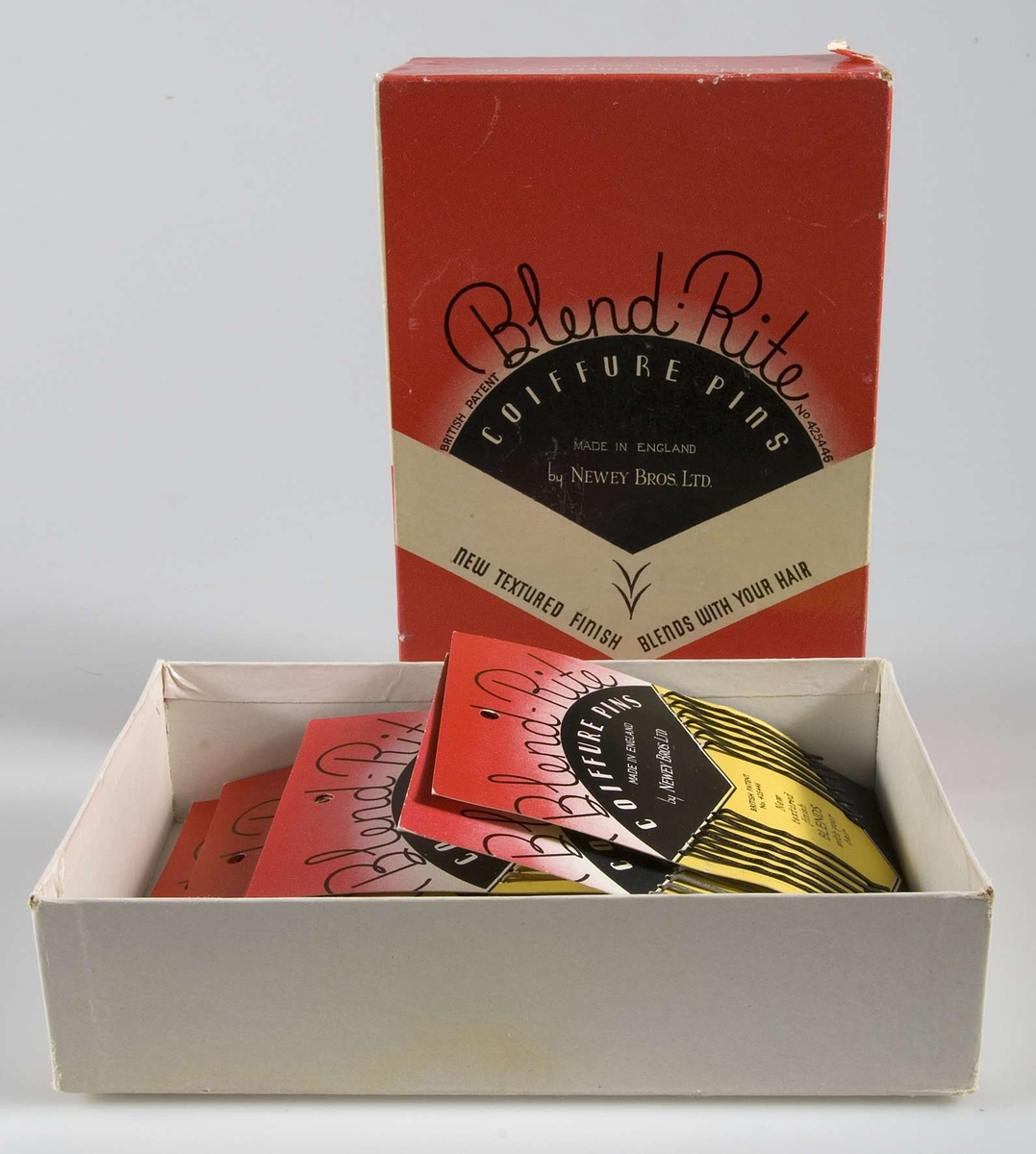Pappask innehållande hårspännen uppsatta på originalpapper. Förpackningen är röd/vit/svart och innehåller 14 papper med 12 smala hårspännen i två olika färger på varje. På asken tryckt text: Blend Rite COIFFURE PINS. På varje papper tryckt 40 öre.