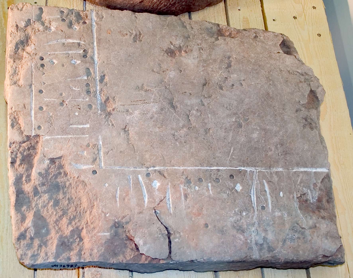 Större fragment av gravhäll, av rödaktig kalksten. Hörndel, två kanter. Kantbård med text, inskriptionen är ifylld med vit färg. Ett flertal hål, cirka 0,5 cm djupa, på kantbården. 