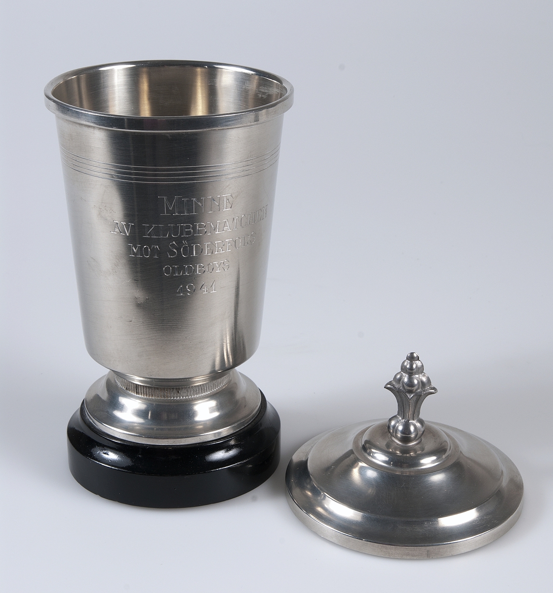 Idrottspris av metall i form av en pokal med lock på underlag av svartmålat trä. På pokalen inskriptionen: MINNE AV KLUBBMATCHEN MOT SÖDERFORS, ODLBOYS 1941.