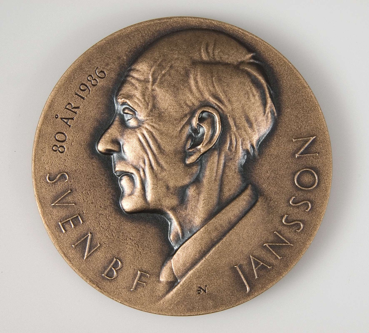 Rund plakett av brons. Tillhörande blå ask. Plakettens motiv på ena sidan: Manshuvud i profil med texten: Sven BF Jansson 80 år 1986. På baksidan: Motiv från runsten. Text: Runforskaren Riksantikvarien Run-Janne.