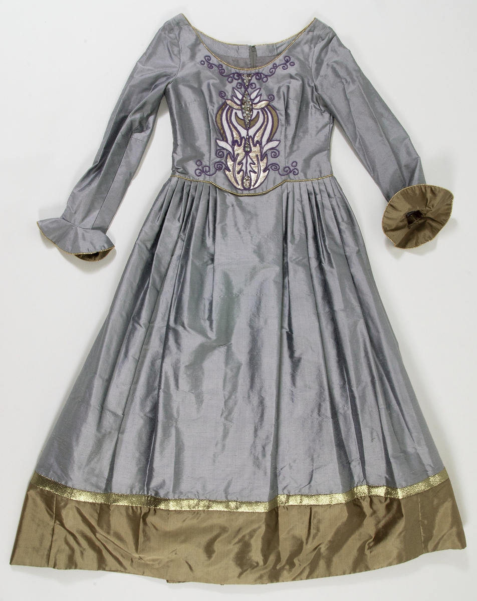 Klänning i grått siden. Figursytt liv. Broderier på framstycket med inslag av pärlor, lila garn och applikationer i guldfärgat och ljusgrått siden. Urringad hals kantad med guldband. På ryggsidan dragkjedja och hyska. Klänningen har smala långa ärmar som är klockade vid handlederna med infodringar av guldfärgat siden, kantat med guldband. Kjolen är utställd och har lagda veck i midjan. Längst ner på klänningen finns en cirka 30 centimeter bred våd av guldfärgat siden. Ovanför våden ett guldfärgat band. Klänningen är fodrad med grått fodertyg.