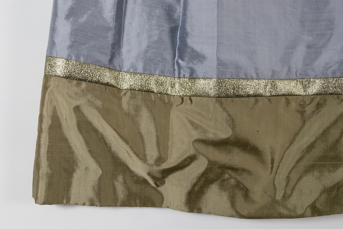 Klänning i grått siden. Figursytt liv. Broderier på framstycket med inslag av pärlor, lila garn och applikationer i guldfärgat och ljusgrått siden. Urringad hals kantad med guldband. På ryggsidan dragkjedja och hyska. Klänningen har smala långa ärmar som är klockade vid handlederna med infodringar av guldfärgat siden, kantat med guldband. Kjolen är utställd och har lagda veck i midjan. Längst ner på klänningen finns en cirka 30 centimeter bred våd av guldfärgat siden. Ovanför våden ett guldfärgat band. Klänningen är fodrad med grått fodertyg.