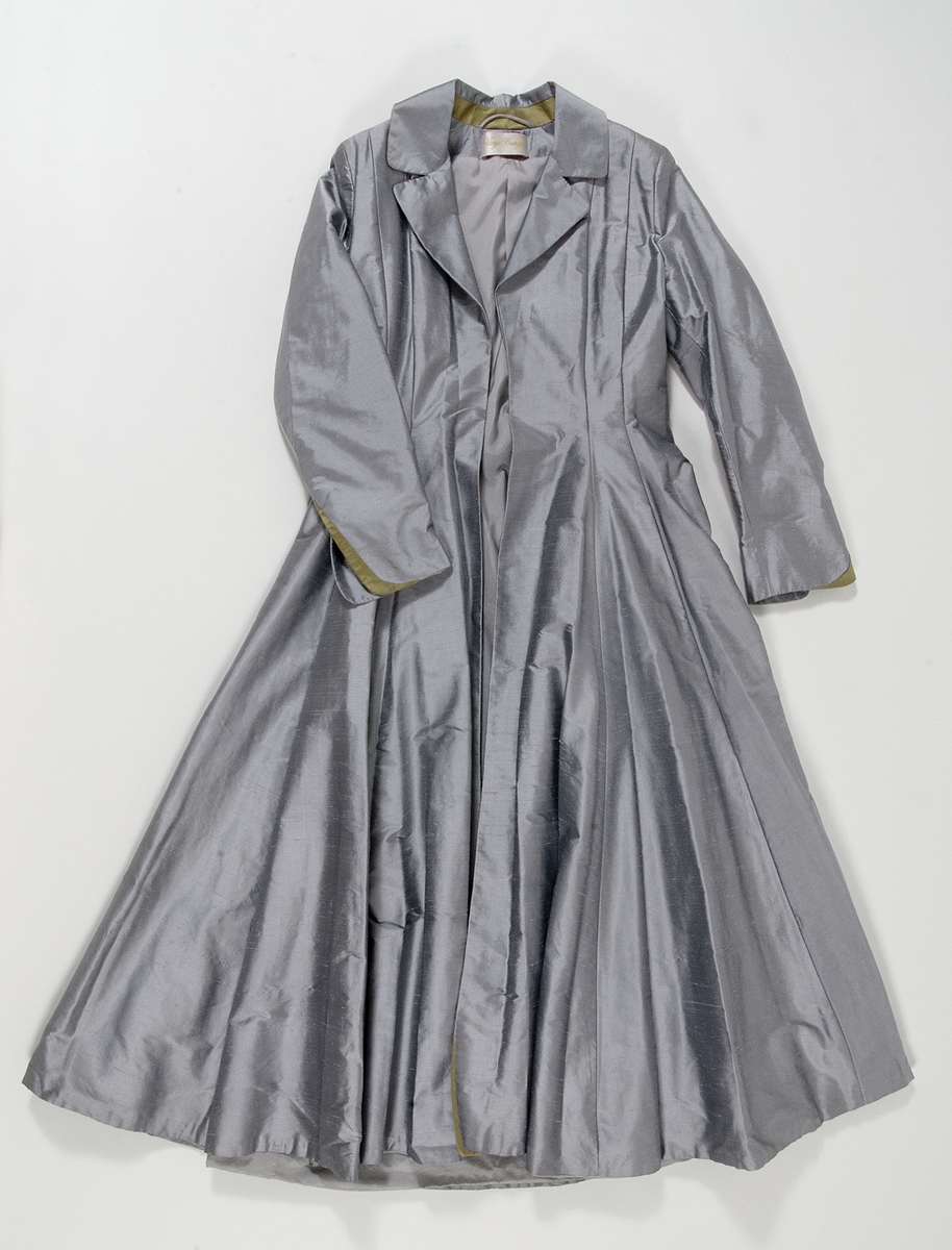 Kappa i grå siden. Kappan, som är formsydd i överstycket, har en utställd kjoldel  med lagda veck runt midjan. Vid halsen en krage med slag. Kappan har långa ärmar med en slits vid handleden. Vid slitsen kant av guldfärgat siden.