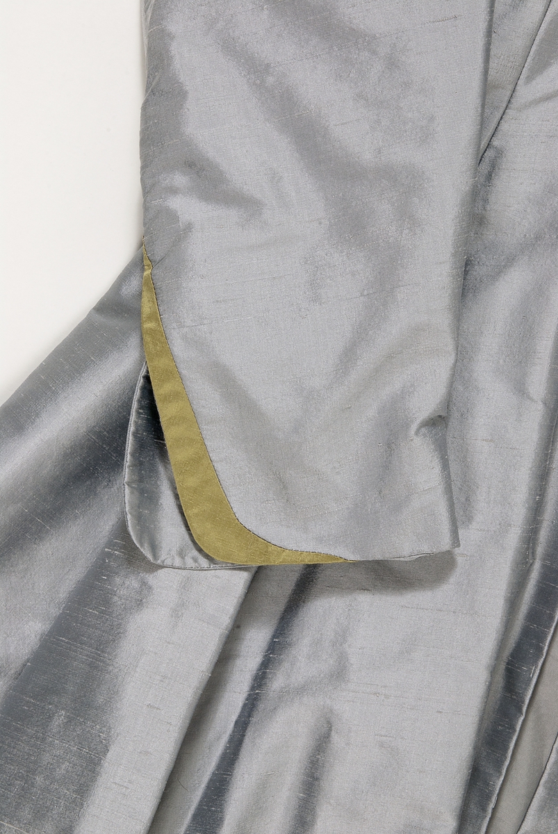 Kappa i grå siden. Kappan, som är formsydd i överstycket, har en utställd kjoldel  med lagda veck runt midjan. Vid halsen en krage med slag. Kappan har långa ärmar med en slits vid handleden. Vid slitsen kant av guldfärgat siden.