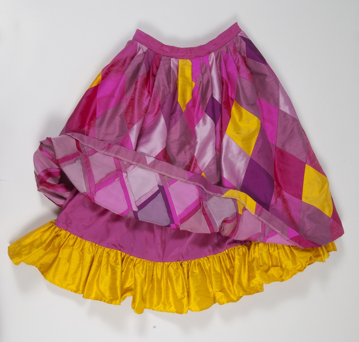 Kjol med rombformade sammansatta bitar i rött, flera nyanser av rosa och gult siden. Kjolen har lagda veck i midjan så att den får vidd. Linning av rosa siden. Kjolen stängs med en rosa knapp och blixtlås på där bak. Kjolen är fodrad med ett rosa fodertyg. Längst ner på fodret finns en 15 cm rynkad våd av gult siden som syns nedanför kjolens kant.