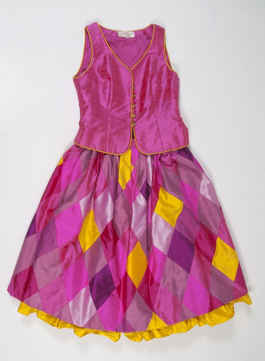 Kjol med rombformade sammansatta bitar i rött, flera nyanser av rosa och gult siden. Kjolen har lagda veck i midjan så att den får vidd. Linning av rosa siden. Kjolen stängs med en rosa knapp och blixtlås på där bak. Kjolen är fodrad med ett rosa fodertyg. Längst ner på fodret finns en 15 cm rynkad våd av gult siden som syns nedanför kjolens kant.