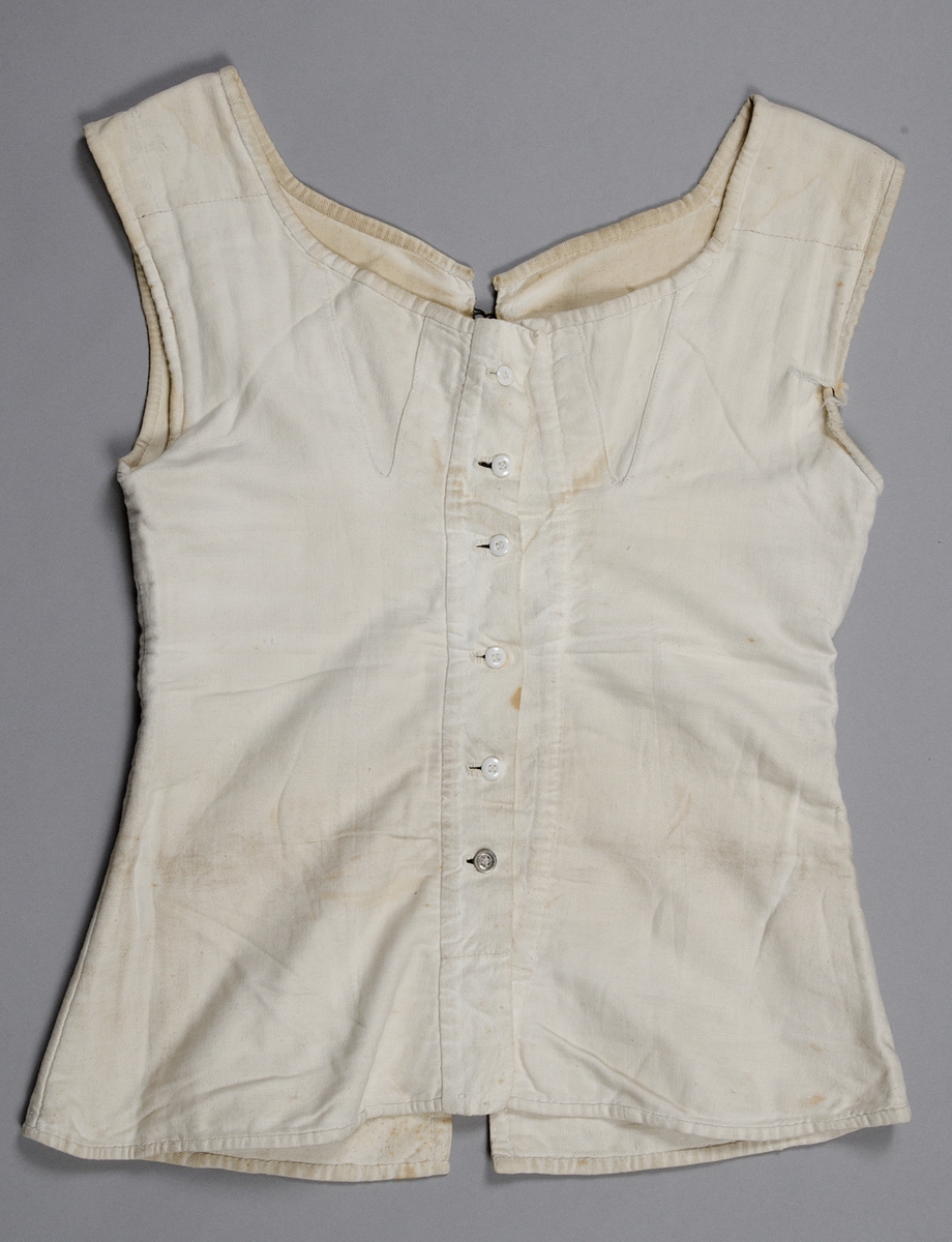 Snörliv, av vit bomull (kypert). Helt handsytt. Två delar som snörs samman bak. Snöret löper genom metallhyskor. Knäpps framtill med sex knappar (fem porslin och en metall). 1840-60-tal (nyrokoko).