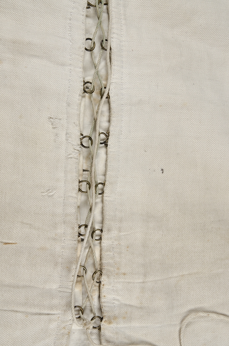 Snörliv, av vit bomull (kypert). Helt handsytt. Två delar som snörs samman bak. Snöret löper genom metallhyskor. Knäpps framtill med sex knappar (fem porslin och en metall). 1840-60-tal (nyrokoko).
