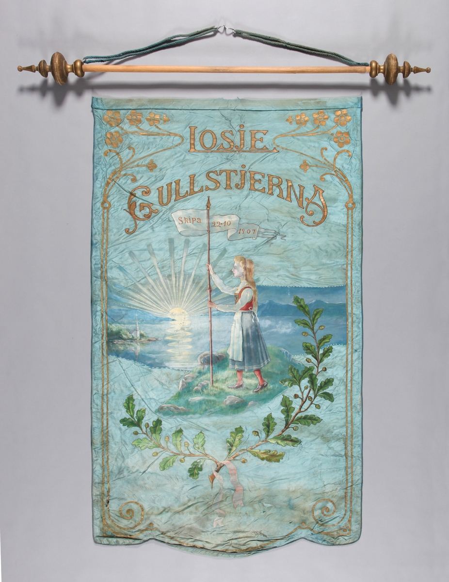 Jente i bunad, vimpel, oppstigande sol, landskap, sjø, eikegreiner med lauv.