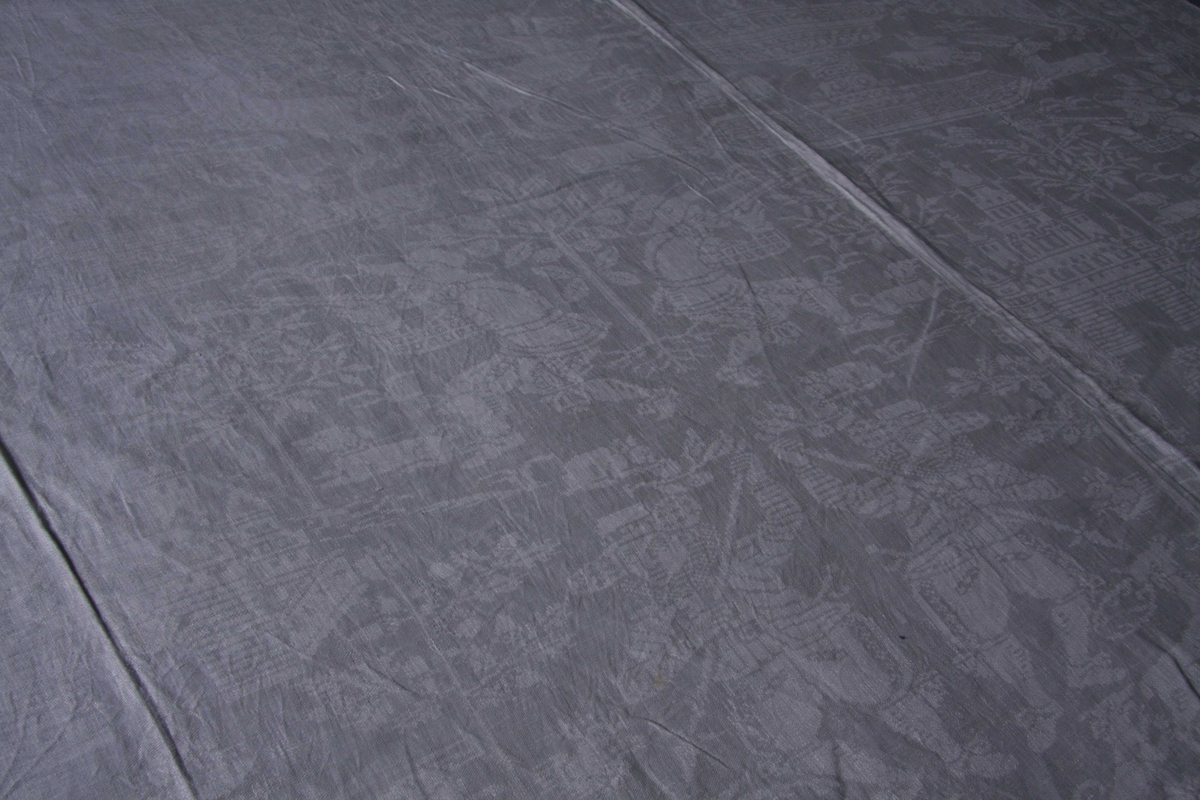 Hvit lindamask med mønster: Enhjørninger flankerer fontene stående på bakbenene med liggende hjerter og andre dyr omkring. Damer til hest med hatt på hodet. Duken har smal fall på kortsidene.