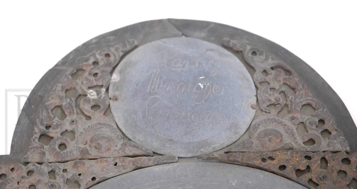 Smidd jernplate påmontert tallskive. Romertall med rester etter bek (fyll). Sekundskive. Øverst et rundt felt med navn på urmaker. Dekorert med støpte messingornamenter. Feltet nederst i venstre hjørne er sekundært, de andre originale. Trolig engelsk importert ornamenter.