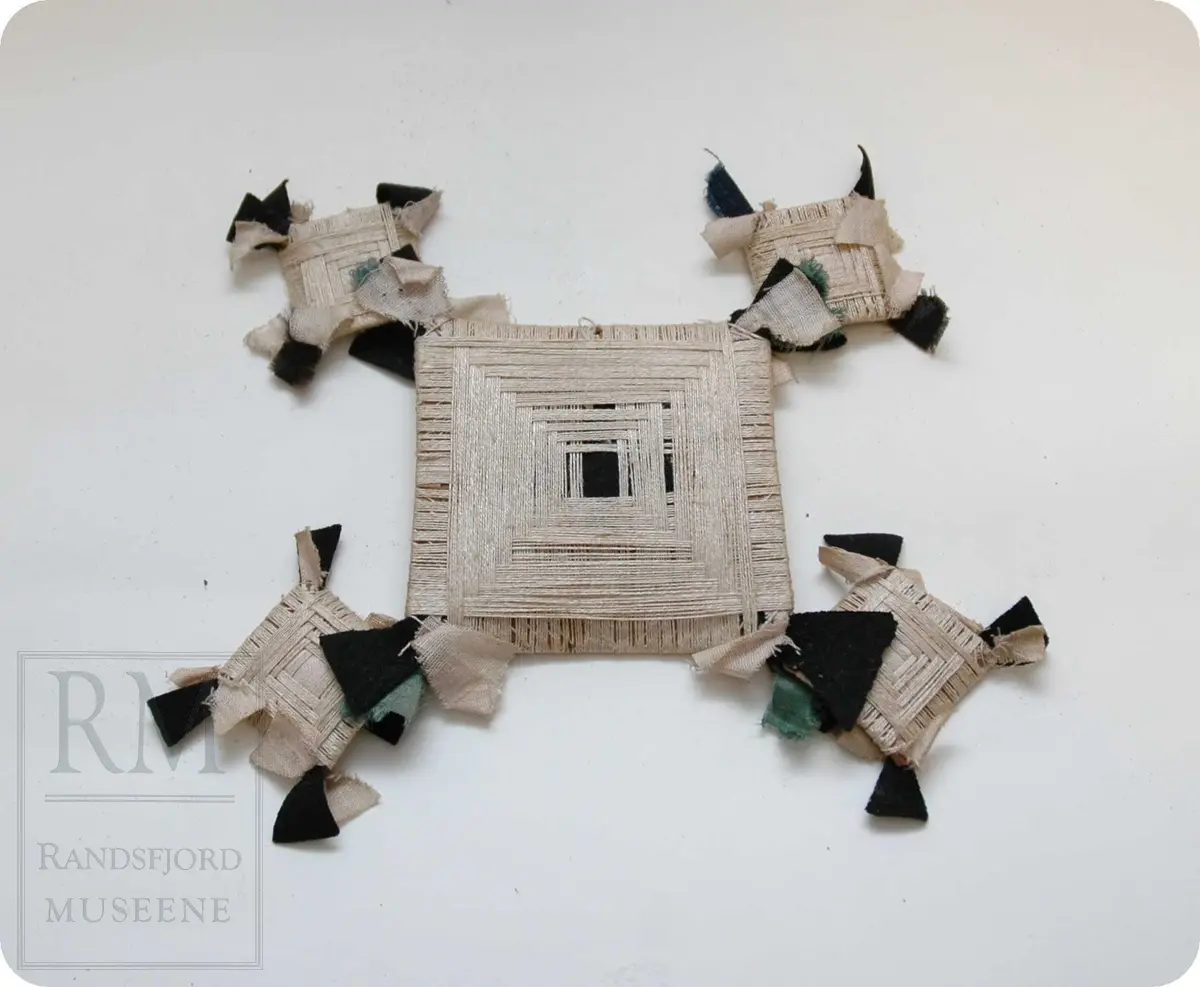 Tråd er surret rundt fem flate pappskiver. En stor og fire små. Stoffbiter er surret fast slik at litt av dem stikker frem under trådenen. De fire små platene er festet til hvert av den store platens hjørner.