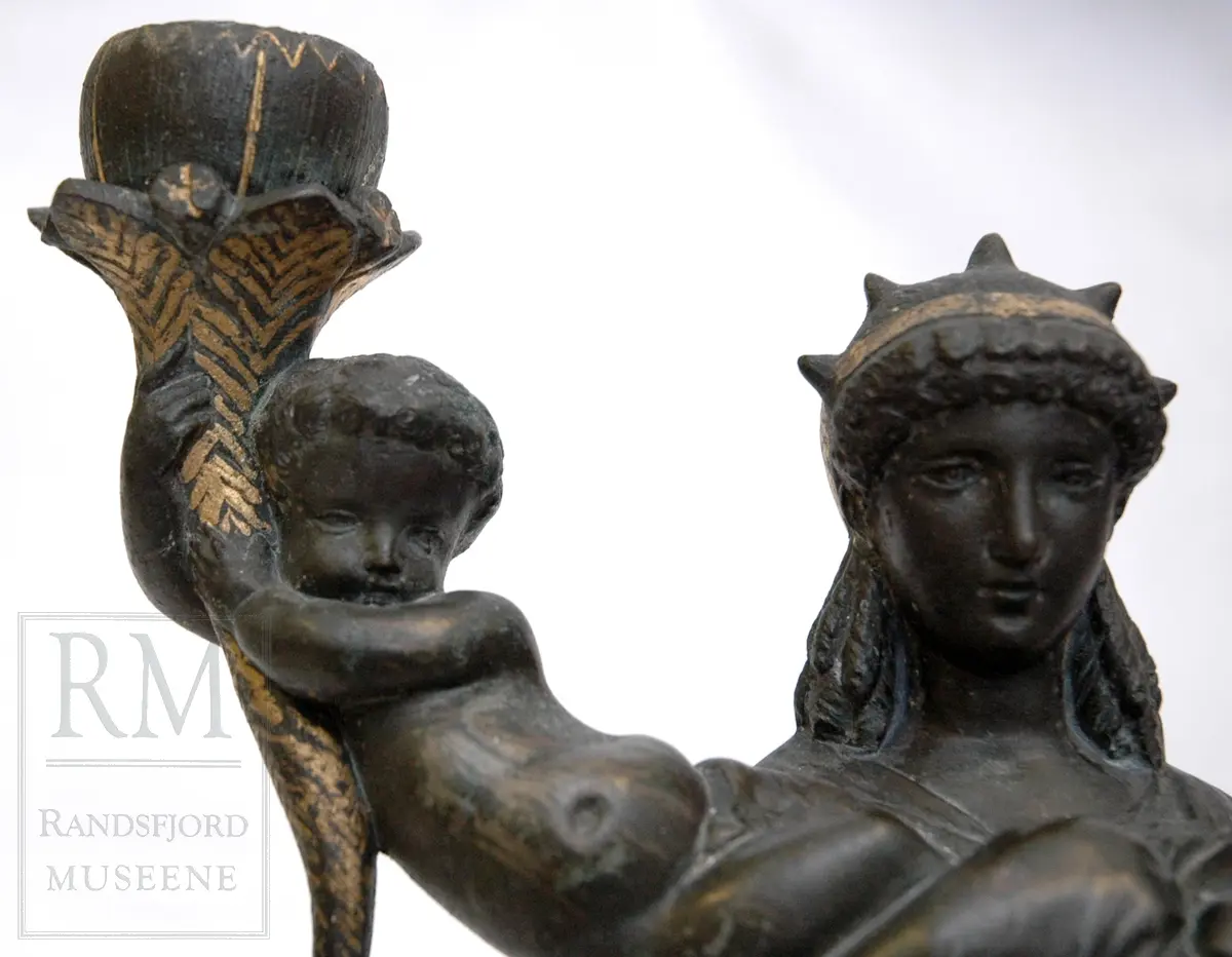 Kvinnefigur på sokkel, med ett barn i hver arm. Barna holder hvert sitt horn (?) med lyseholder. Noe forgylling. Løse lysmansjetter. b) mangler sokkel.