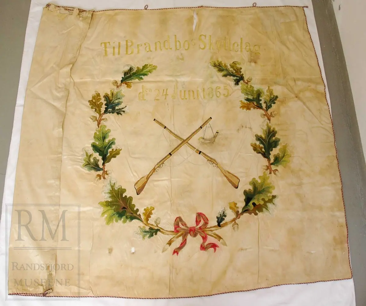 Fane av hvit silke. Løpegang på den ene siden har revnet. Brodert tekst, samt eikebladskrans med sløyfe omkring to gevær med påhengende krutthorn.