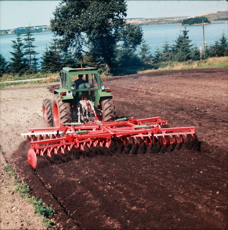 Stor 48-skålers skålharv i arbeid bak en Deutz-traktor, Svein Sigmundstad i traktoren, i bakgrunnen Frøylandsvatnet